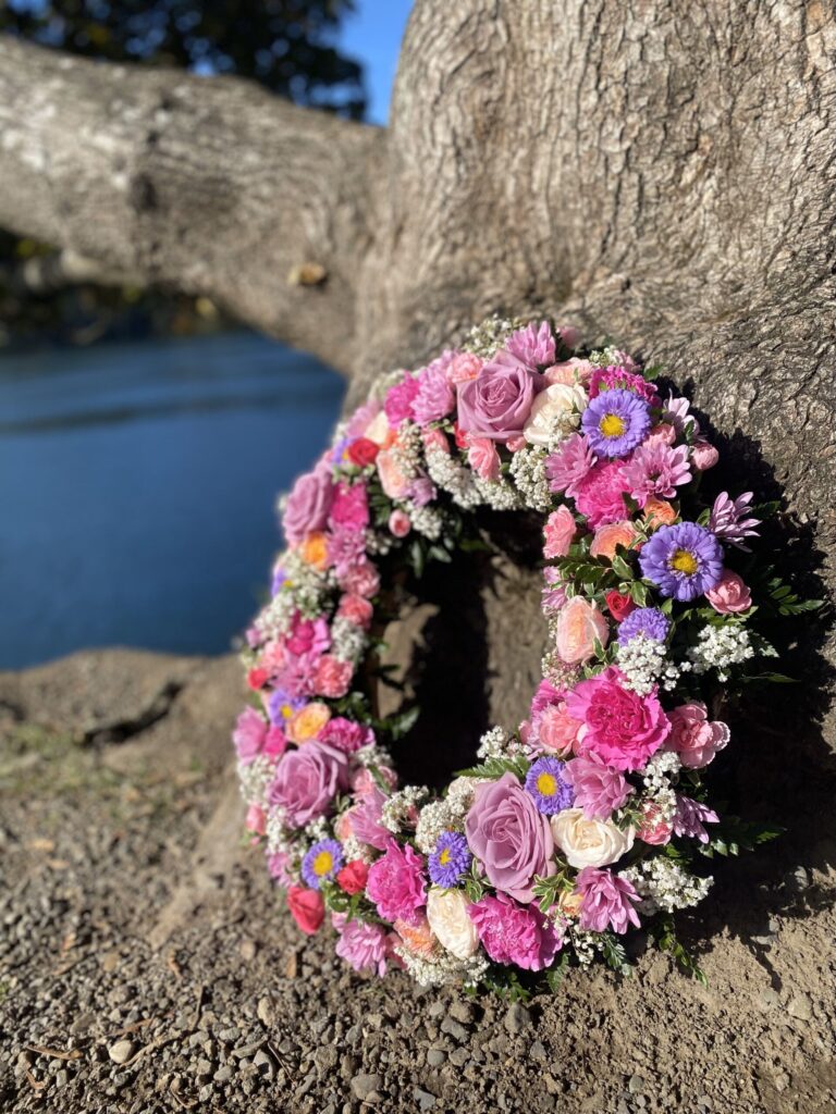 A vibrant floral wreath with pink, purple, and white flowers rests against the base of a large tree, symbolizing remembrance and the legacy of whole body donation.
