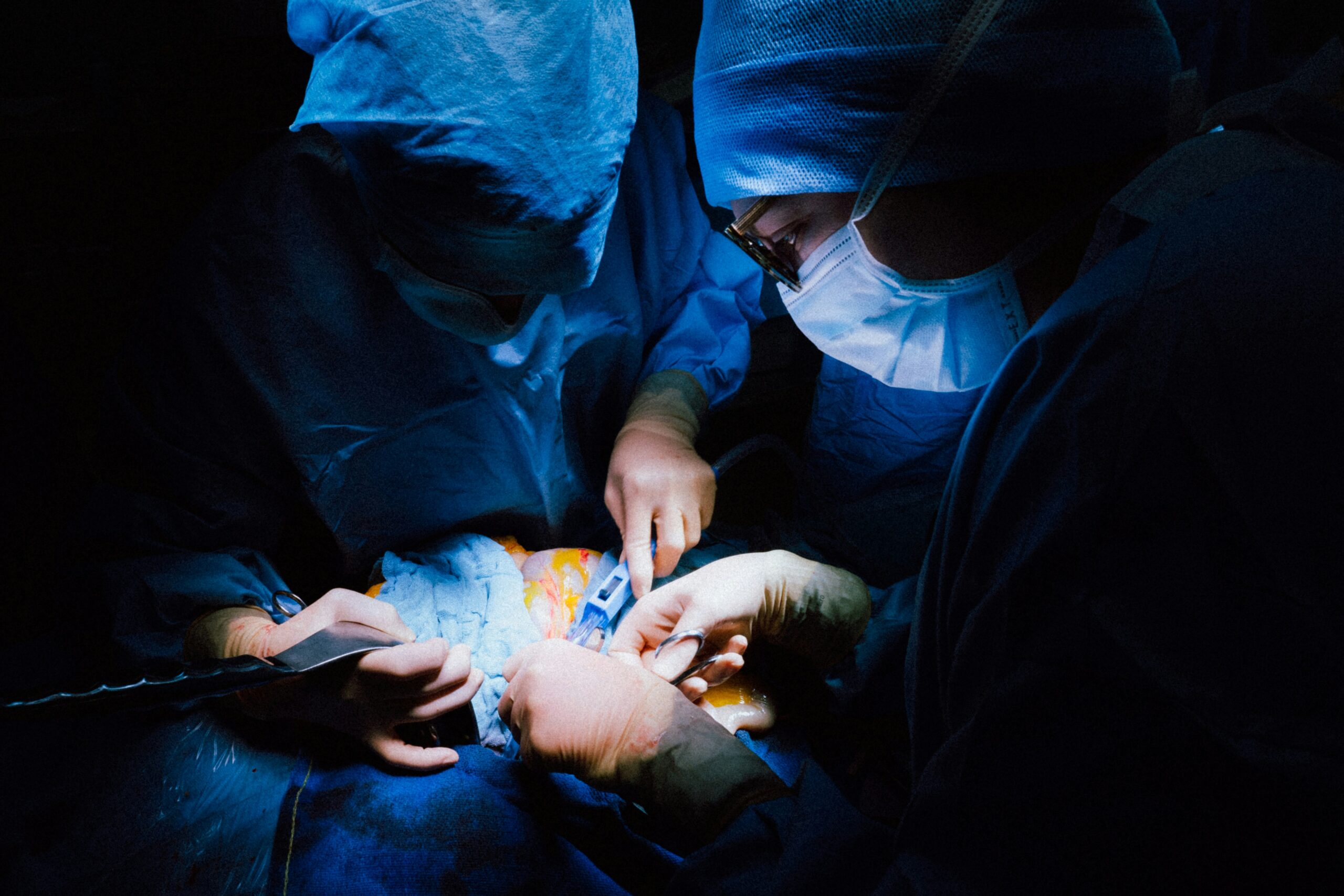 Surgeons in a dimly lit operating room performing a surgical procedure, illuminated by bright, focused lights, wearing blue surgical gowns, masks, and gloves.
